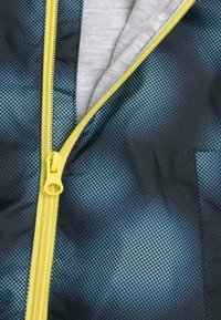 Veste zippée avec un motif à pois bleu et noir, dotée d'une fermeture éclair jaune et d'une doublure intérieure grise. Comprend une poche patch.