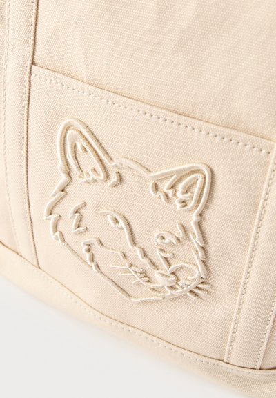 Ύφασμα κρεμ με ραμμένη περίγραμμα του προσώπου μιας αλεπούς σε μια τσέπη, που δείχνει την υφή και τις καθαρές ραφές.