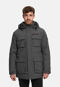 Homme portant une veste d'hiver grise à capuche avec quatre poches à rabat à l'avant, debout devant un fond clair uni.