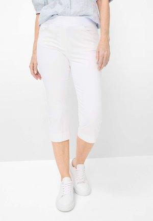 Kvinde iført hvide korte bukser, lyseblå mønstret bluse og hvide sneakers, stående på en hvid baggrund.