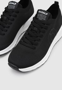 Zapatillas de malla negras con cordones negros resistentes y suela blanca. Presentan una puntera redondeada y una superficie texturizada con un diseño de logo sutil.
