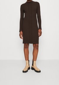 Personne portant une robe à manches longues côtelée de couleur marron foncé, longueur genou, avec des bottines beiges épaisses sur un fond blanc.