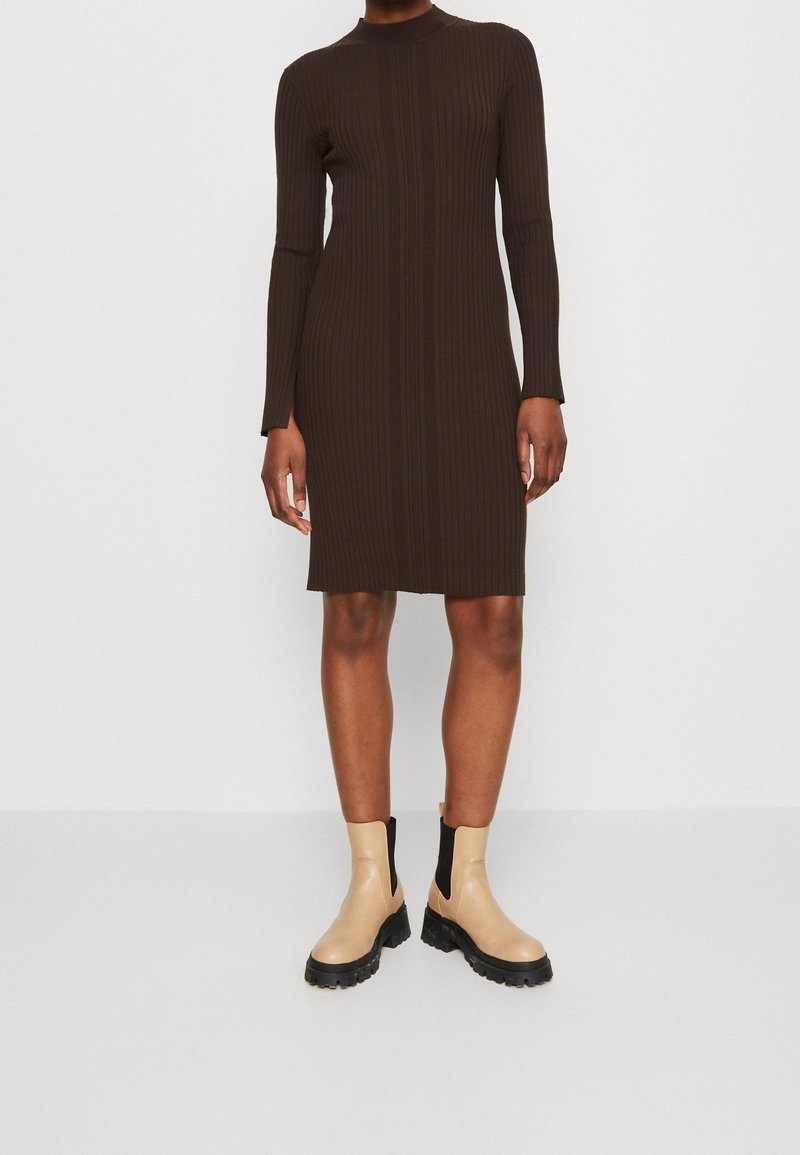 Personne portant une robe à manches longues côtelée de couleur marron foncé, longueur genou, avec des bottines beiges épaisses sur un fond blanc.