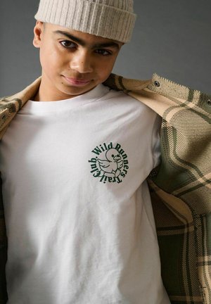 Jeune personne portant un bonnet en tricot beige et une veste à carreaux sur une chemise blanche avec un logo de canard et le texte "Wild Runners Trail Club".