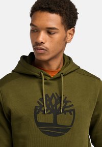 Felpa verde oliva realizzata in un morbido tessuto texturizzato. Presenta un logo di un albero nero al centro, con i lacci sul cappuccio.
