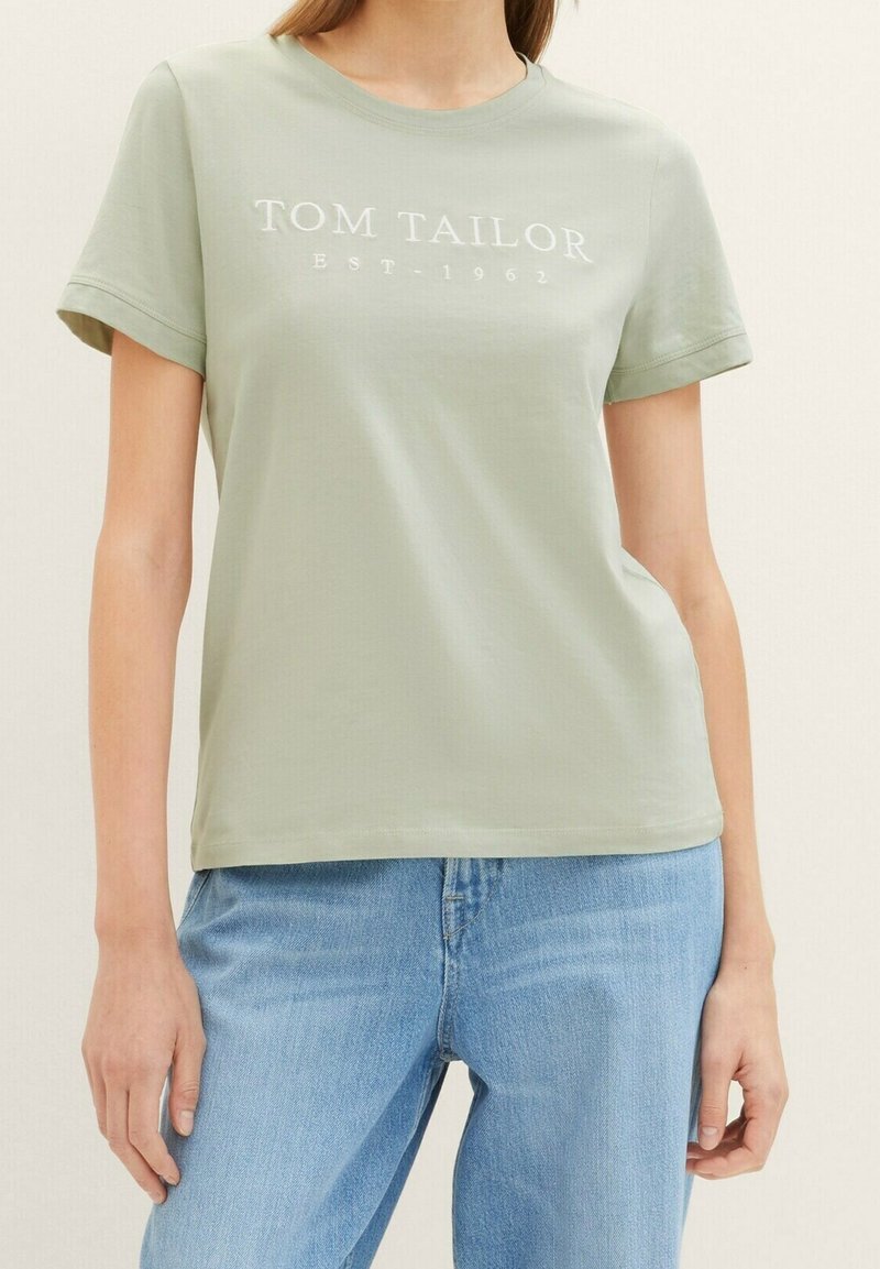 Lys grøn bomulds T-shirt med korte ærmer, med hvid trykt tekst "TOM TAILOR EST 1962." Simpel rund halsudskæring, afslappet pasform.