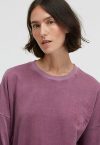 Lila Sweatshirt mit einer runden, strukturierten Grafik mit der Aufschrift "SENSES" auf der Brust. Verfügt über einen Rundhalsausschnitt und überschnittene Schultern, hergestellt aus weichem Stoff.