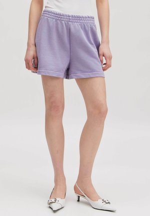 Person trägt fliederfarbene Fleece-Shorts und weiße Slingback-Pumps mit niedrigem Kittenheel auf hellem Hintergrund.
