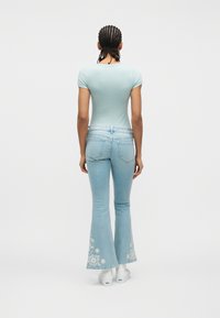 Άτομο με πλεξούδες που φοράει στενό ανοιχτό μπλε t-shirt και ανοιχτό μπλε τζιν με φουντωτά μπατζάκια, διακοσμημένα στο κάτω μέρος με λευκή κεντητή ανθοδέσμη, καθώς και λευκά αθλητικά παπούτσια.