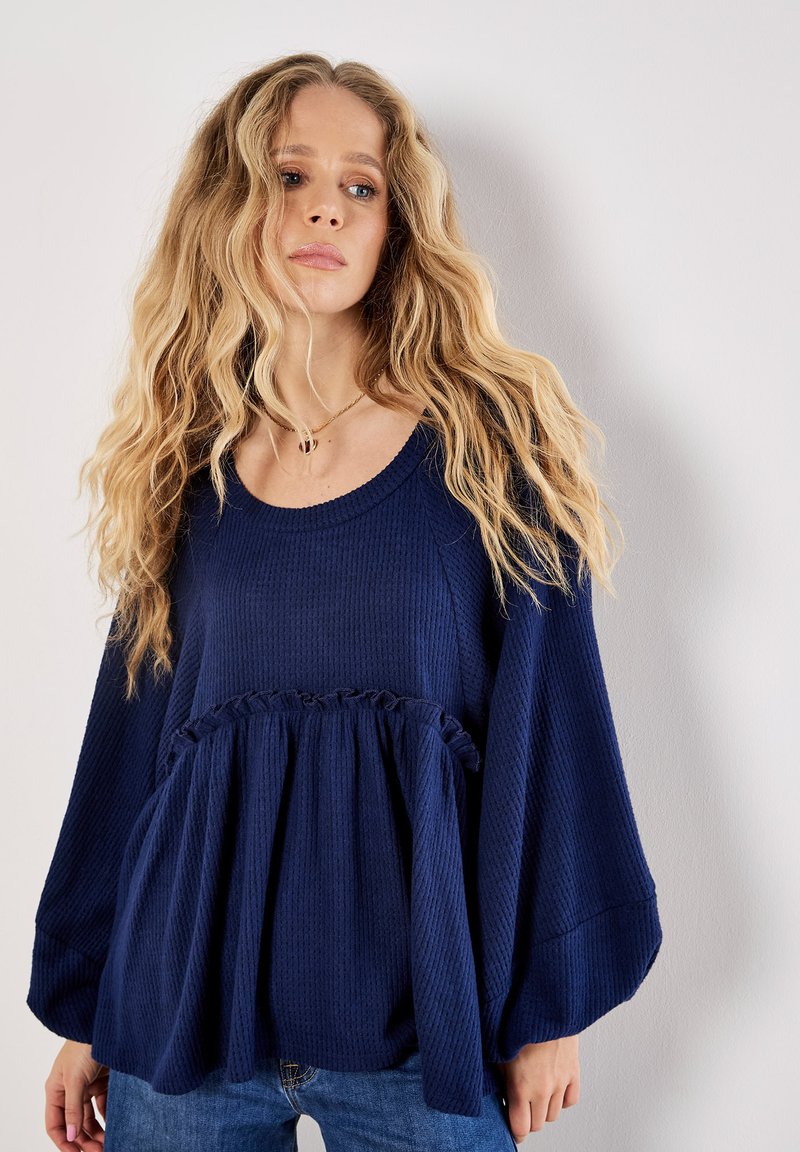 Navyblaues, strukturiertes Oberteil mit Rundhalsausschnitt, langen Ärmeln und gerafftem Taillendetail, kombiniert mit lässigen Jeans.