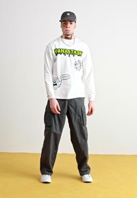 Valkoinen pitkähiha shirt, jossa on vihreät grafiikat ja mustat sarjakuvakoristeet, yhdistettynä tummaan cargo-housuun ja valkoisiin tennareihin.