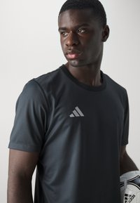 Sort trænings T-shirt med korte ærmer og rund hals, med et gråt Adidas-logo på den øverste venstre bryst. Holder en fodbold.