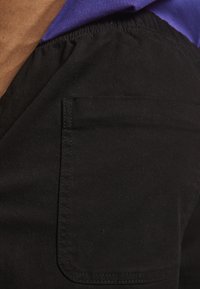 Schwarze Canvas-Hose mit sichtbar engem Seitentasche, die eine glatte Textur und subtile Nähdetails aufweisen. Getragen mit einem lila Hemd.