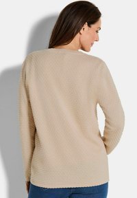 Beige Langarmpullover mit strukturierten, erhabenen Punkten, runder Ausschnitt und lockerer Passform, kombiniert mit blauen Jeans.