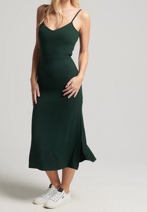 Mujer con un vestido midi sin mangas de color verde oscuro combinado con zapatillas blancas, de pie frente a un fondo liso de color gris claro.