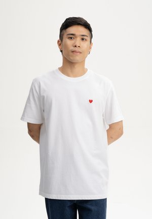 Jonge man met kort donker haar en oorbellen, gekleed in een wit T-shirt met een klein rood hartje op de linkerborst, staand tegen een effen achtergrond.