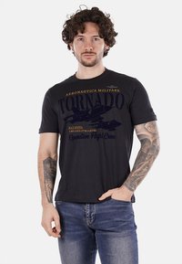 Camiseta de algodón negra con gráfico de un jet y texto, cuello redondo, mangas cortas y un corte clásico. Tatuaje visible en el antebrazo izquierdo.