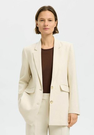 Světle béžový blazer s klopami, dvěma předními kapsami a jedním knoflíkem, nošený přes tmavohnědý žebrovaný top.