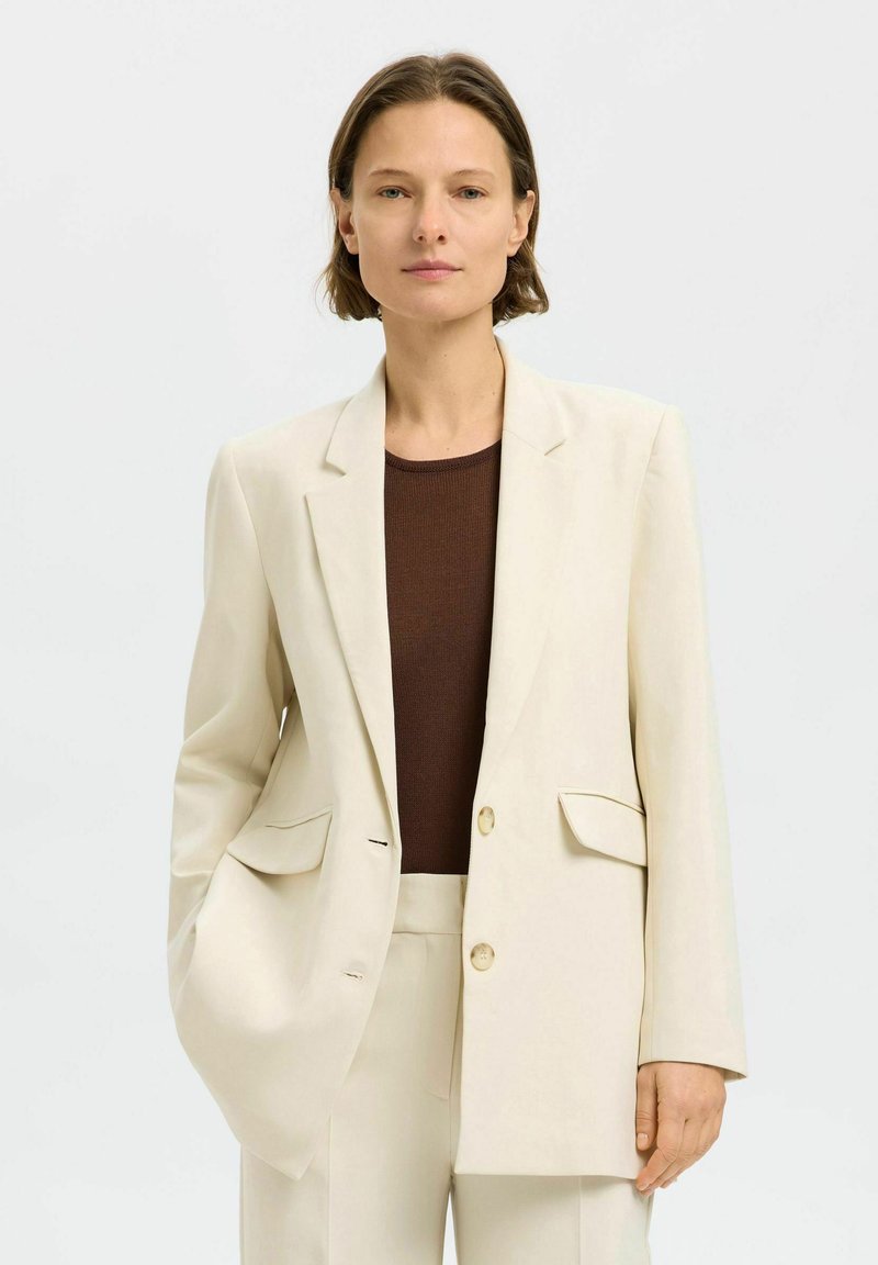 Lys beige blazer med spidse klapper, to frontlommer og en enkelt knaplukning, båret over en mørkebrun ribbet top.