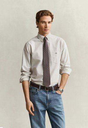 Uomo con camicia a righe abbottonata, cravatta fantasia, jeans blu, cintura intrecciata e orologio al polso sta in piedi con una mano in tasca.