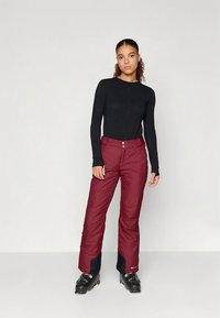 Bordeauxrode skibroek met een gladde textuur, taps toelopende pasvorm en verstelbare tailleband; gecombineerd met een zwart shirt met lange mouwen en skistiefels.