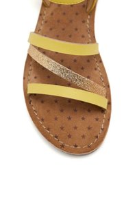 Sandalia amarilla con correas, que presenta acentos dorados brillantes, una plantilla acolchada marrón con un patrón de estrellas y costuras detalladas a lo largo de los bordes.