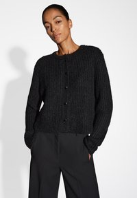 Schwarze Strickjacke mit geripptem Muster, Rundhalsausschnitt und schwarzen Knöpfen. Kombiniert mit schwarzen, weit geschnittenen Hosen.