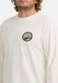 Mann trägt ein elfenbeinfarbenes Langarmshirt mit kleinem bunten Billabong-Abenteuerlogo auf der linken Brust.