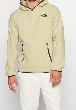 Homme portant une polaire à capuche North Face beige clair avec bordure noire et poches avant zippées, associée à un pantalon blanc.