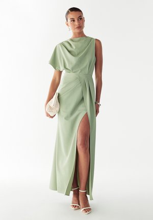 Abito lungo verde con top drappeggiato asimmetrico e spacco laterale. Tessuto liscio, vita aderente, abbinato a una borsa bianca testurizzata.