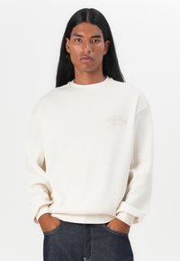 Cremfarbener, gerippter Sweatshirt mit rundem Halsausschnitt, überschnittenen Schultern und Bündchenärmel. Dezentes Logo-Detail auf der Brust.