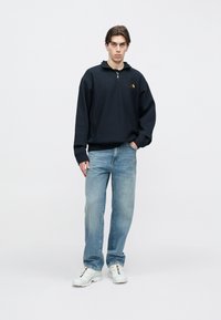 Μπλε σκούρο φούτερ με φερμουάρ και μικρό λογότυπο στο στήθος, σε συνδυασμό με ανοιχτόχρωμο τζιν και λευκά αθλητικά παπούτσια. Απλός σχεδιασμός και άνετη εφαρμογή.