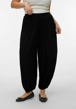 Kvinne iført løstsittende svarte bukser med elastisk midje og rynkede buksekant, kombinert med svarte og hvite ballettsko med mønster.