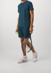 Tyrkysové sportovní tričko a šortky, lehký materiál, tenisová raketa v ruce, drží žlutý tenisový míček, bílé boty Nike s černými akcenty.