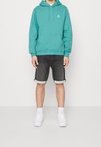 Sudadera con capucha verde azulado y cordones, con un bolsillo tipo canguro; combinada con pantalones cortos de mezclilla oscuros con dobladillos enrollados; zapatillas deportivas blancas.