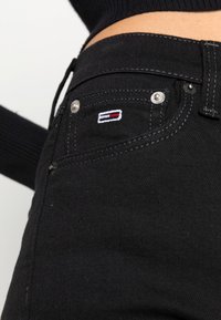 Jeans de mezclilla negra con una vista en primer plano del bolsillo delantero que presenta herrajes en plata y un pequeño logo bordado en rojo, blanco y azul.