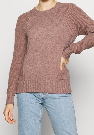 Kvinde iført en lilla ribstrikket sweater med kabeldetaljer på ærmer og sider, kombineret med lyseblå jeans, hånd på hoften.
