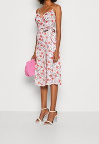 Robe midi fleurie avec des fleurs roses et orange sur un fond clair. Dotée d'un design portefeuille, de fines bretelles et d'une taille nouée, portée avec des talons blancs.