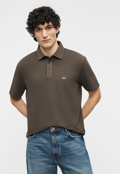 Jeune homme aux cheveux bouclés portant un polo marron avec le logo "A|X" et un jean bleu, debout les mains derrière le dos sur un fond blanc.