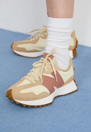 Person iført beige og cremefarvede sneakers med lyserødt "N" logo og hvide sokker, der står på blåt tæppe.