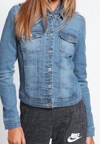 Chaqueta de mezclilla azul claro con mangas largas, cierre de botones, dos bolsillos en el pecho y costuras sutiles. Llevada sobre pantalones de chándal gris oscuro.