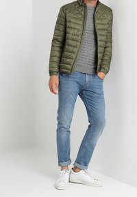 Olivenfarvet, quiltet jakke med ståkrave, grå sweater, lyseblå jeans og hvide sneakers. Oprullede ærmer på jeansene.