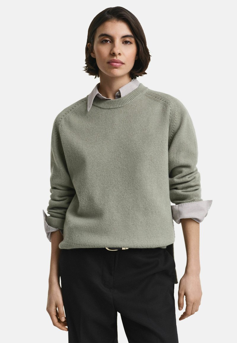 Donna con capelli scuri e corti, che indossa un maglione a maglia grigio chiaro sopra una camicia con colletto, pantaloni neri, in piedi di fronte a uno sfondo bianco.