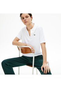 Vit polo-tröja med krage, försedd med en grön Lacoste-logga, kombinerad med mörkgröna byxor. Slät tyg och figursydd design.