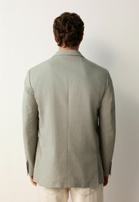 Lys grå skræddersyet blazer lavet af et glat stof, med en enkelt slids bagpå og knapmanchetter, parret med lyse bukser.