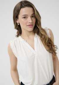 Blouse blanche à manches courtes avec des bords volants, un décolleté en V partiel et un tissu drapant lisse. Associée à un jean en denim noir.