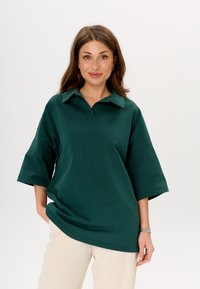 Donkergroene, oversized polo-shirt met een kraag en korte mouwen, gemaakt van een zachte stof. Heeft een relaxte pasvorm en minimale details.