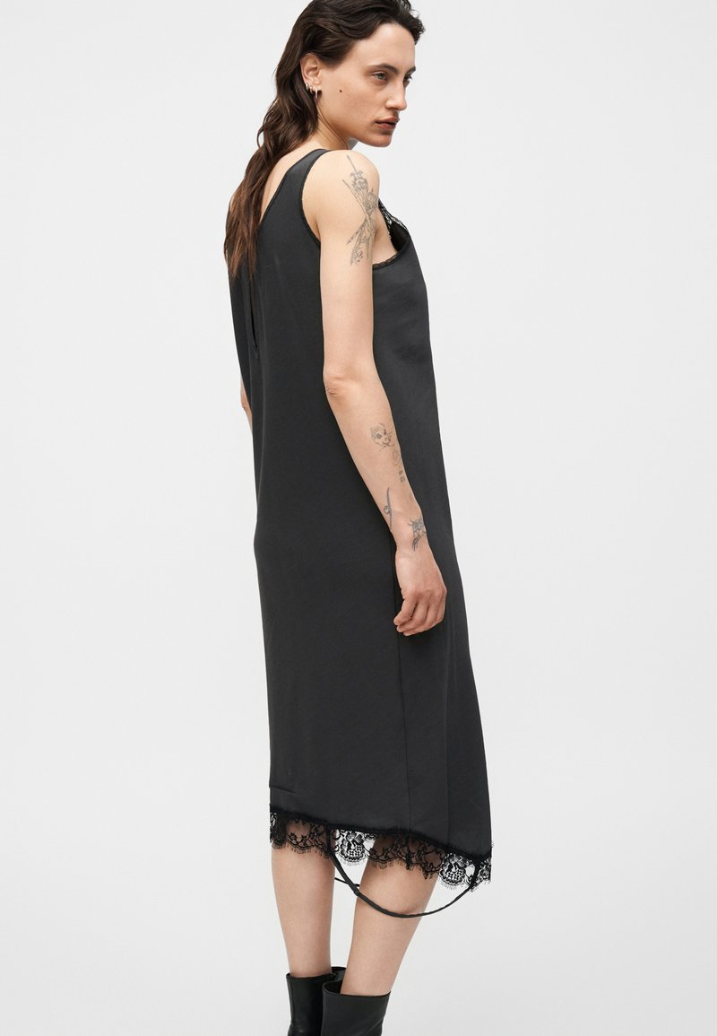 Frau mit Tattoos trägt ein ärmelloses schwarzes Midikleid mit Spitzenbesatz und schwarze Stiefeletten, steht im Profil vor einem einfarbigen Hintergrund.