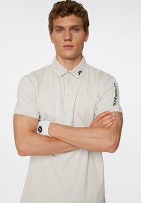 Beige kortärmad polotröja med knäppning, krage och svarta logodetaljer på kragen och ärmen; mjuk tygstruktur.