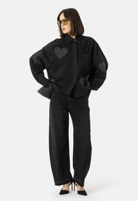 Žena v nadměrné černé denimové košili s nášivkami srdcí, širokých černých džínách, černých vysokých podpatcích, nese malou pletenou černou kabelku.